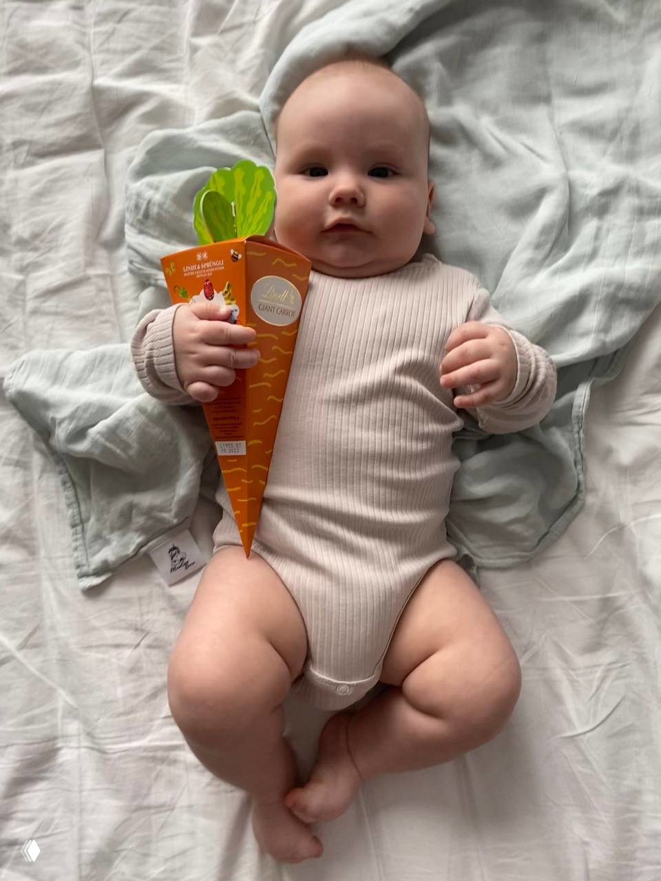 Грудной ребёнок в бежевом боди держит конус с конфетами в виде морковки, снимок в домашней уютной обстановке с мягкими тканями на фоне.