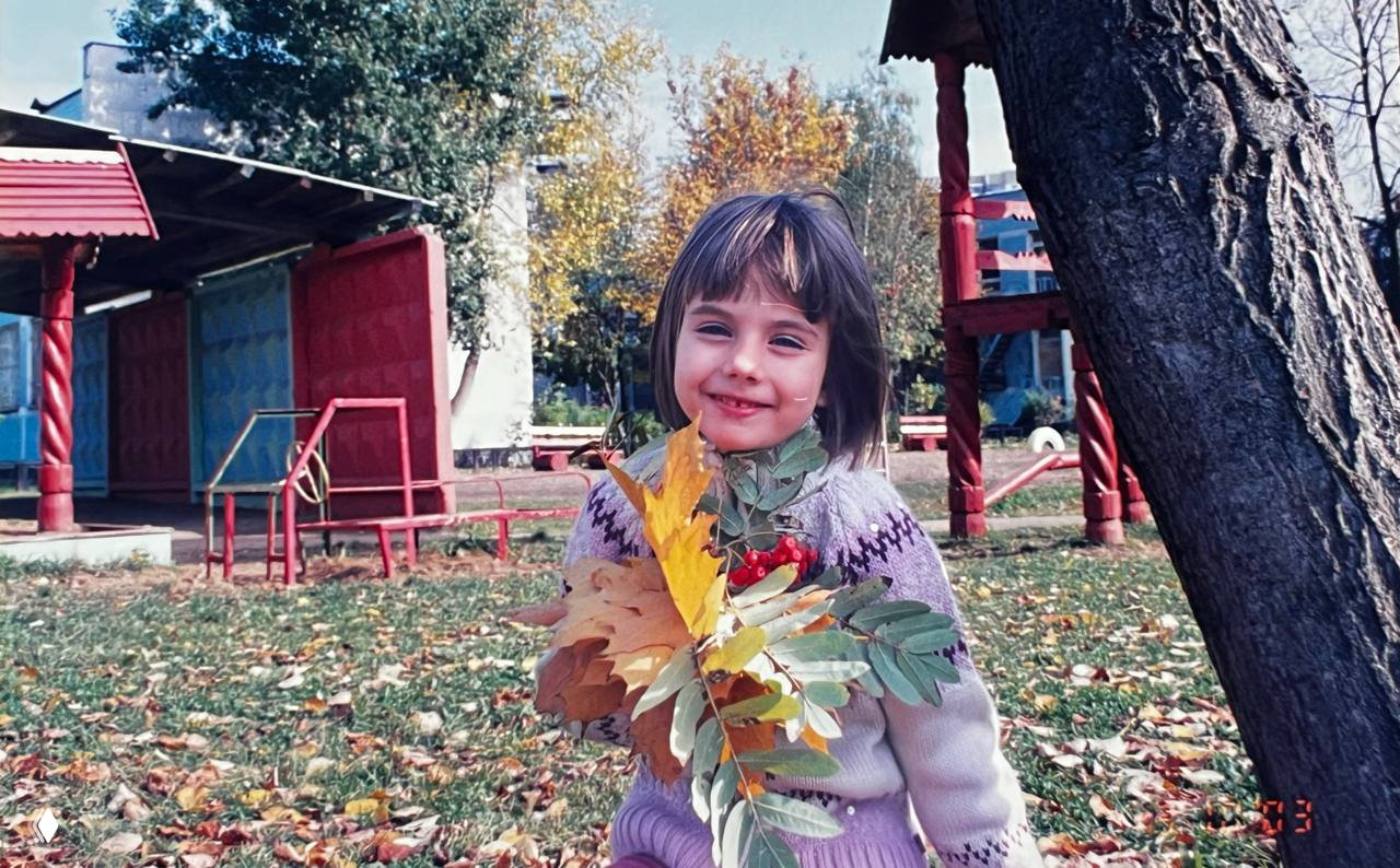 Осенняя фотография на детской площадке: девочка с веткой и жёлтыми листьями на фоне горок и деревьев — ностальгический кадр.