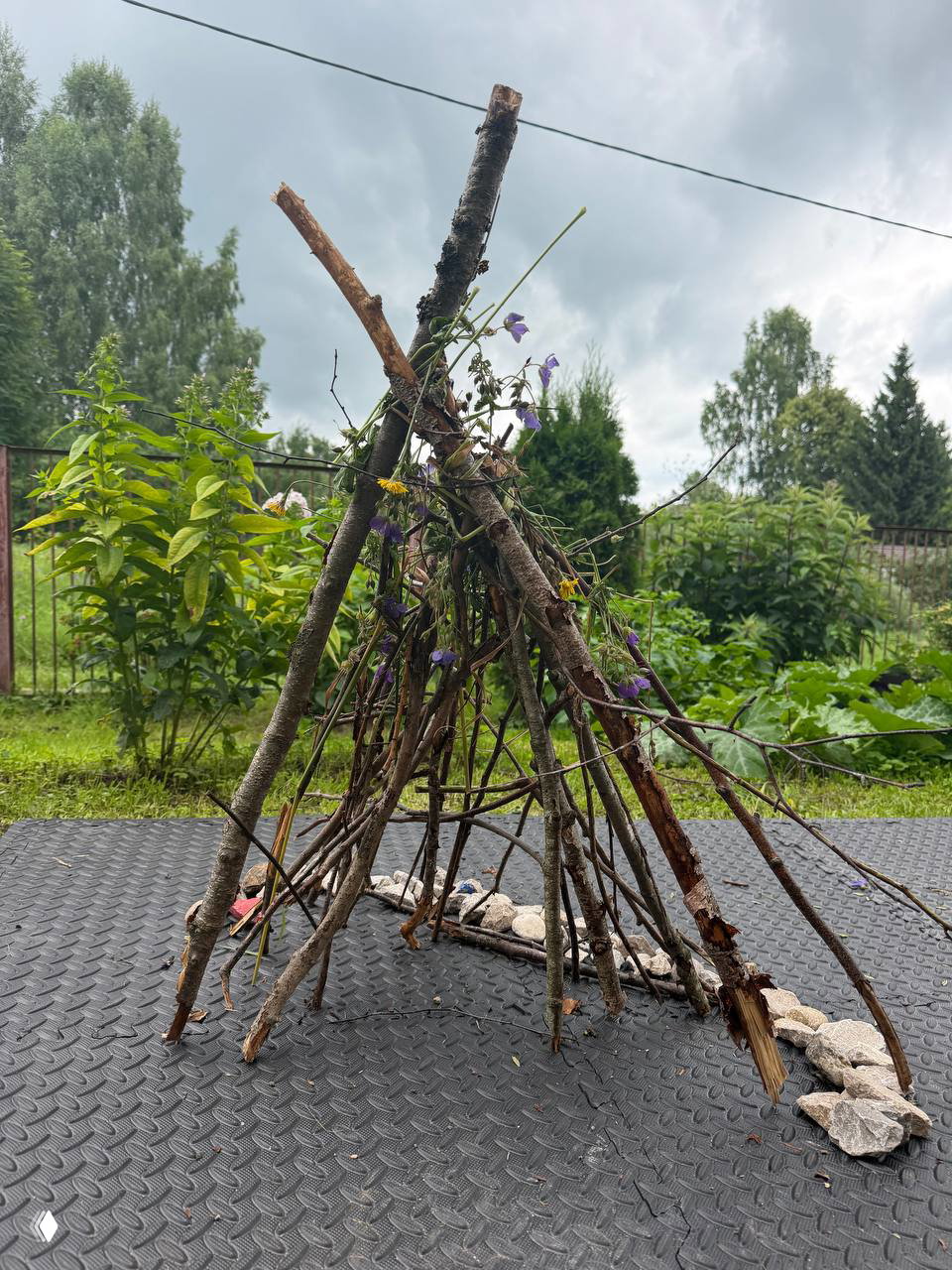Низкая конструкция из палочек, собранная в виде шалаша, украшенная цветами и окружённая полосой из камней — почти готовый мини‑дом для букашек.