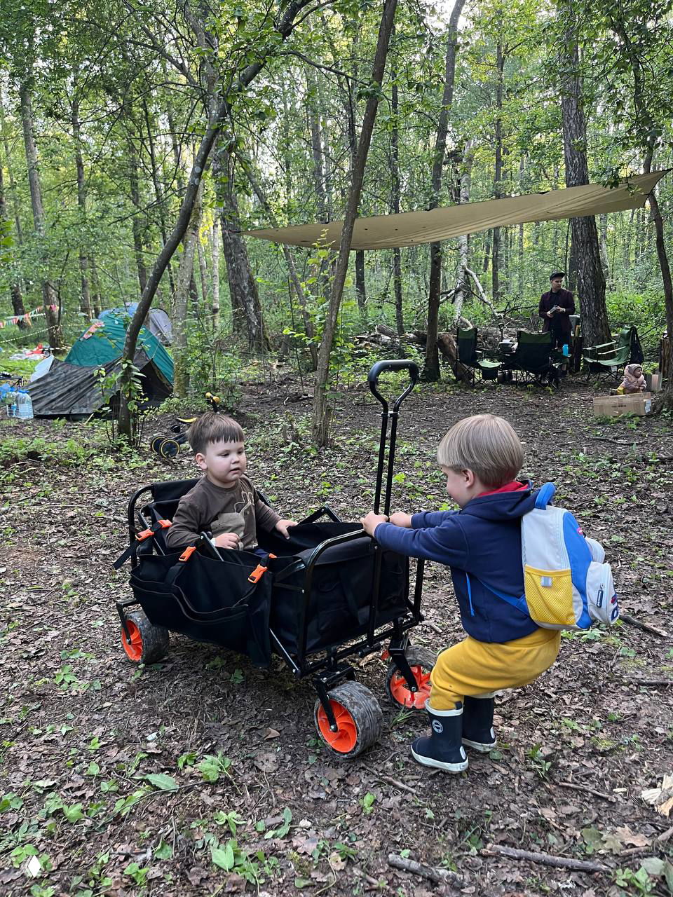 Дети играют у складной тележки-вагончика на лесной стоянке, на заднем плане тенты и столовая зона лагеря с подвешенным тентом.