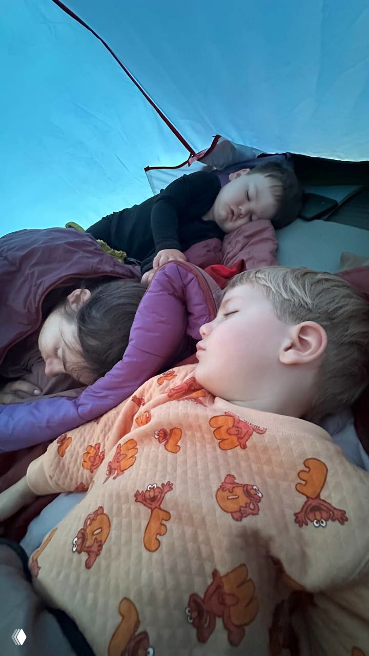 Спящие дети и взрослый внутри палатки на надувных ковриках и в спальниках — тёплая сцена ночёвки в палаточном лагере для семей.