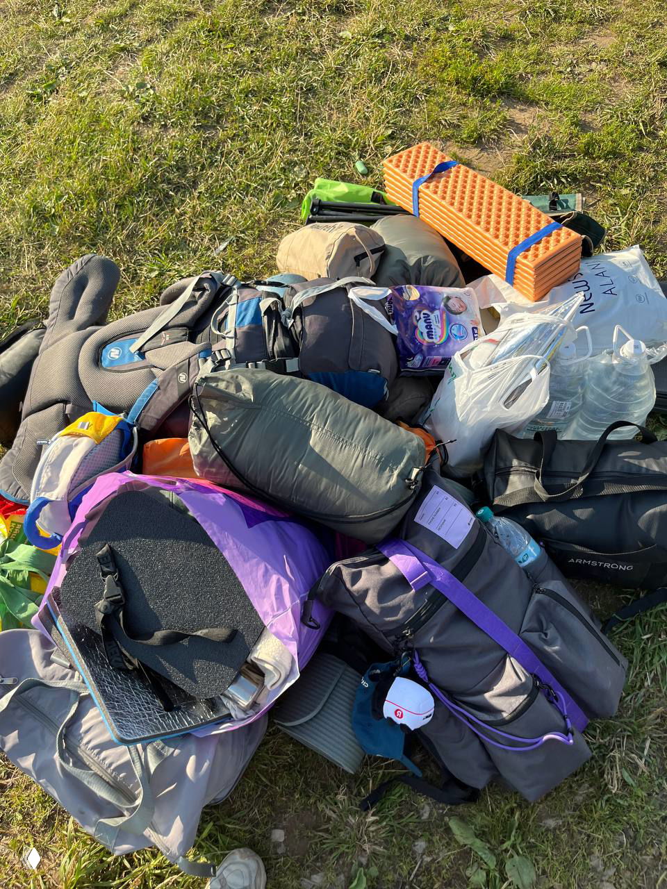 Куча походного снаряжения: рюкзаки, коврики, спальники и пакеты с водой на траве — визуализация объёма вещей для выезда с ребёнком.