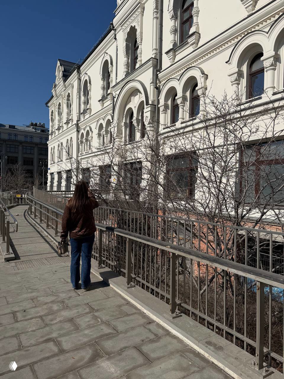 Женщина идёт по тротуару вдоль белого исторического здания и фотографирует кусты с ранними почками; городской весенний пейзаж.