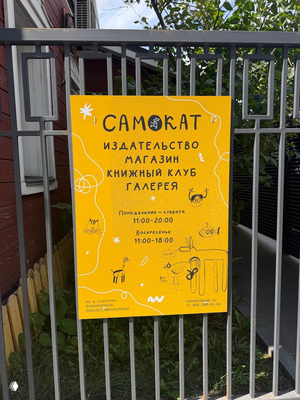 Жёлтый постер издательства «Самокат» на ограде с режимом работы и названием, уличный кадр рядом с входом в книжный клуб.
