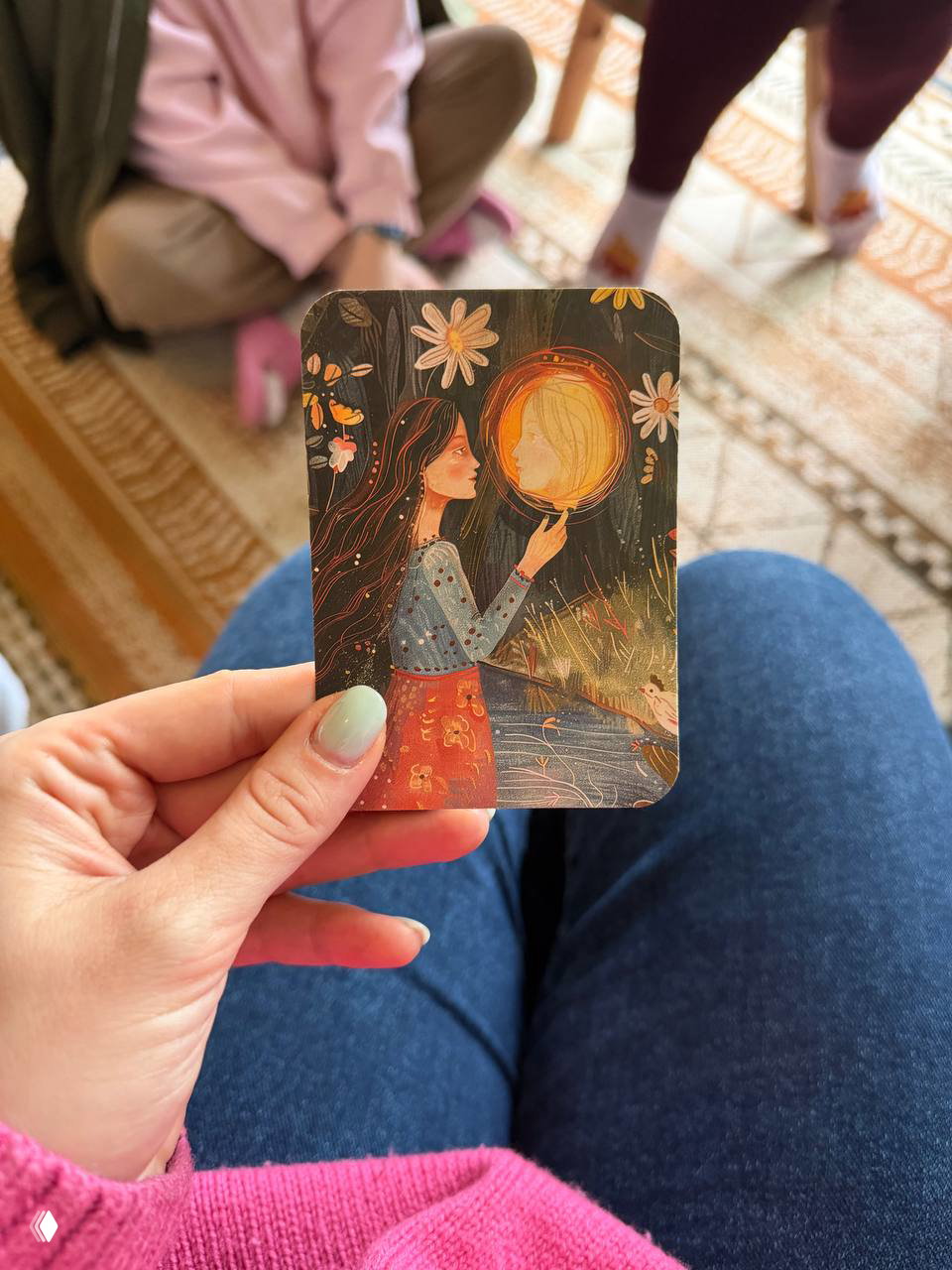 Рука взрослого держит маленькую иллюстрированную карточку с тёплым рисунком; фон — ноги и ковер, спокойный момент отдыха и небольшая игра для родителей.