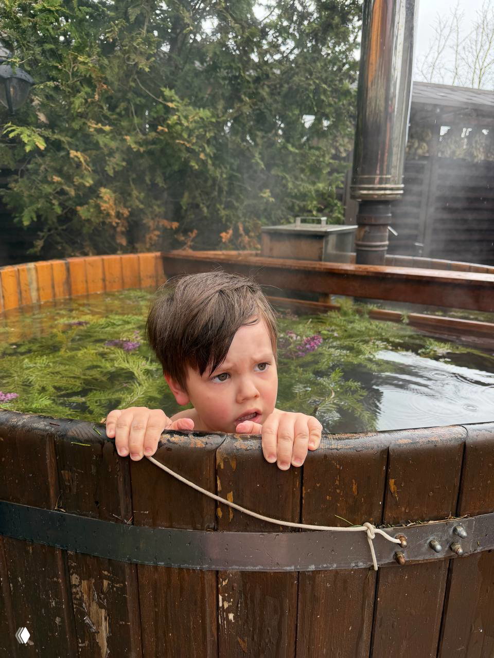 Ребёнок в горячем деревянном чанe держится за край, виден пар и плавающая растительность в воде — момент релаксации в семейной бане.