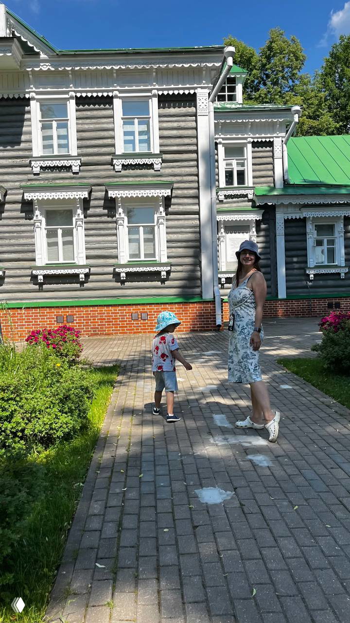 Мама и ребёнок идут по мощёной дорожке возле резного деревянного дома-музея с зелёной крышей и белыми наличниками, летний солнечный день.
