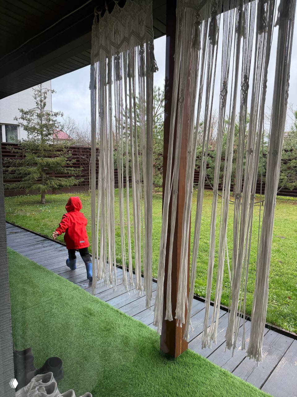 Вид с веранды на зелёный двор: ребёнок в красной куртке идёт по настилу, по бокам висят длинные макраме-занавесы — уютная уличная зона комплекса.