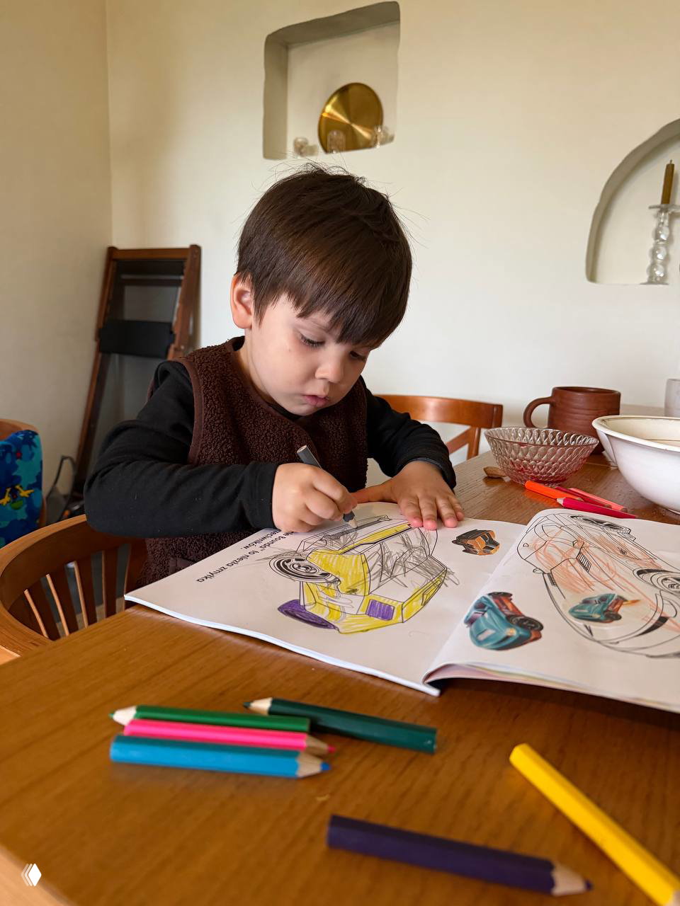 Ребёнок раскрашивает раскраску за деревянным столом в зоне отдыха: цветные карандаши и спокойная атмосфера для занятий внутри комплекса.