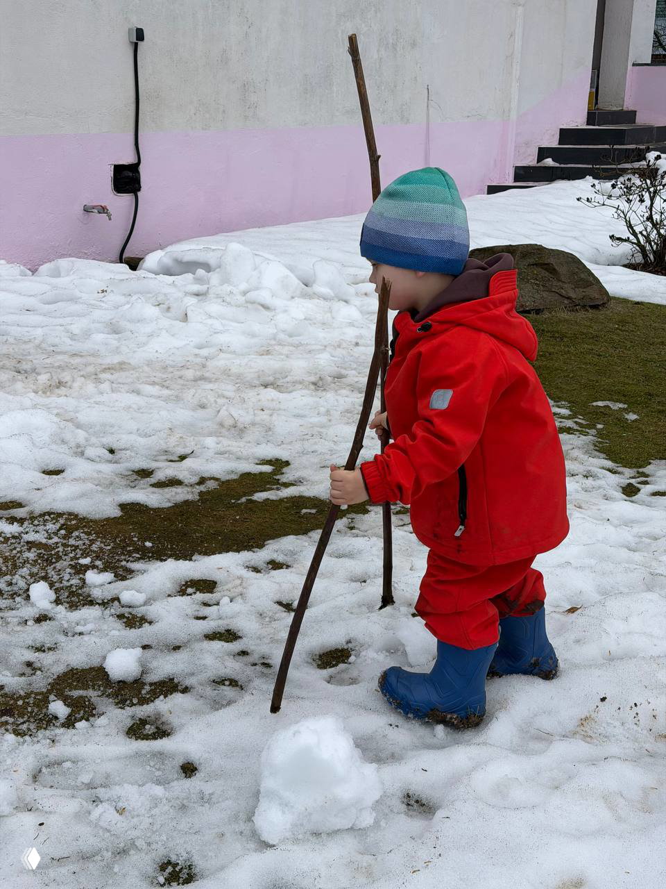 Маленький ребёнок в красном комбинезоне и тёплой шапке несёт две тонкие палки по тающему снегу на газоне рядом с домом — начало дачного сезона и игры на улице.