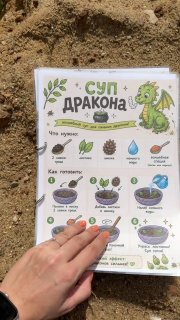 Иллюстрированный лист с рецептом «Суп дракона» на песке для детской грязевой кухни; в кадре частично видна детская рука.