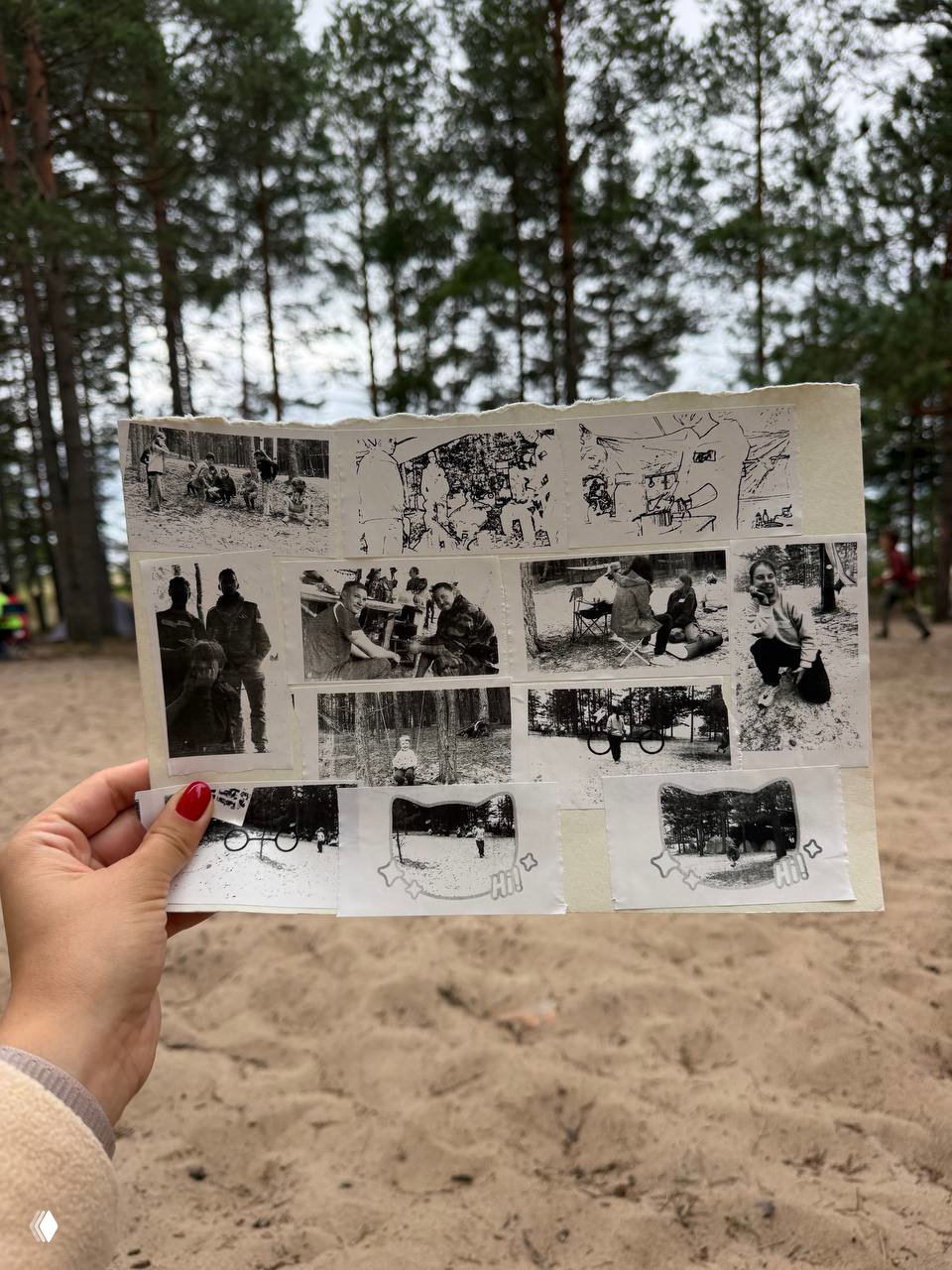 Рукa с красным маникюром держит лист с отпечатанными черно-белыми фотографиями лагерной жизни на фоне песочного поля и сосен.