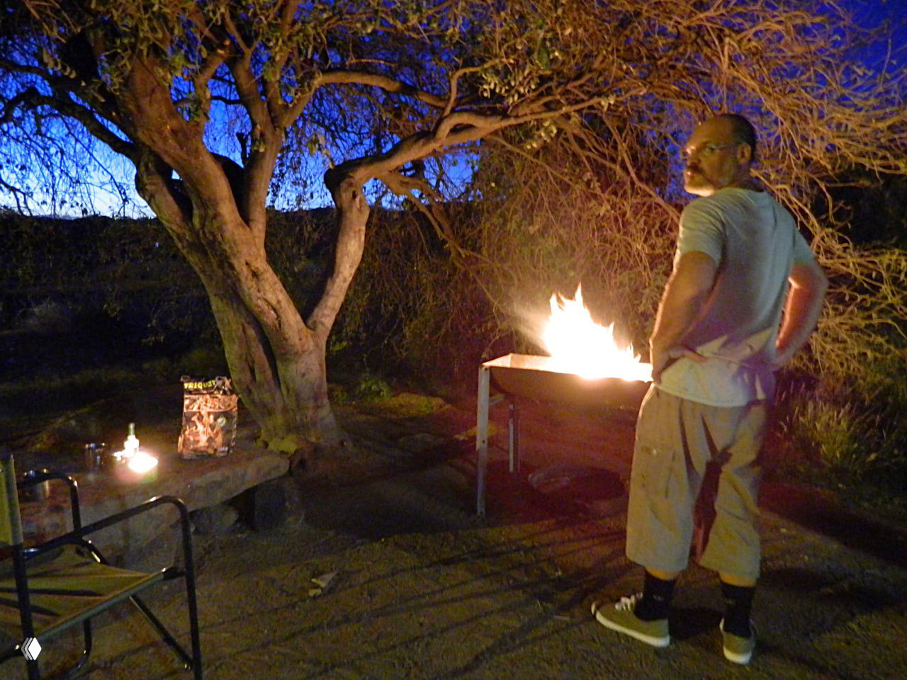 Вечерний кадр у костра в кемпинге: огонь на гриле, стол и мужчина рядом — атмосфера ночёвки на дикой стоянке с возможностью приготовления еды.