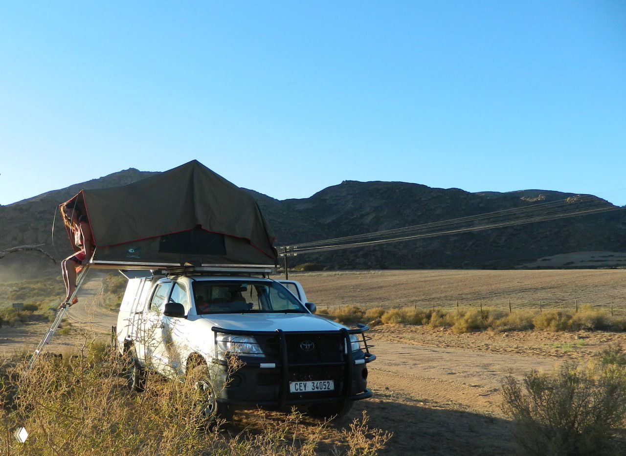 Белый джип с палаткой на крыше в открытом ландшафте с холмами на заднем плане — пример мобильного автокемпинга по африканским дорогам.