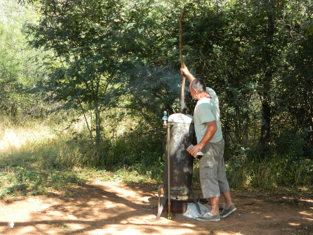 Мужчина протапливает бойлер-«самовар» в кустах рядом с кемпингом, чтобы получить горячую воду — пример простых инженерных решений в удалённых стоянках.