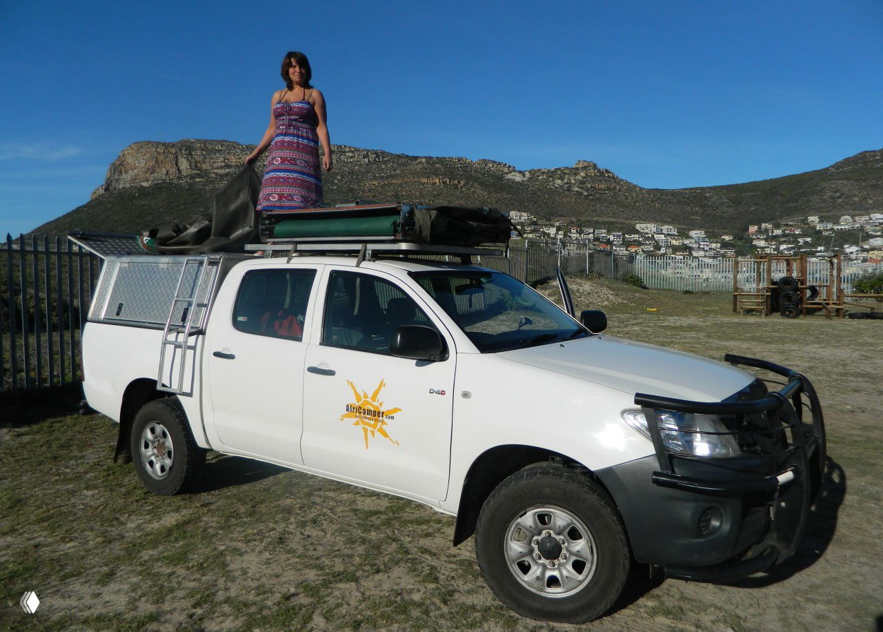 Женщина стоит на крыше белого пикапа с установленной палаткой, вокруг виднеется прибрежный город и горы — сцена подготовки к ночёвке на машине.