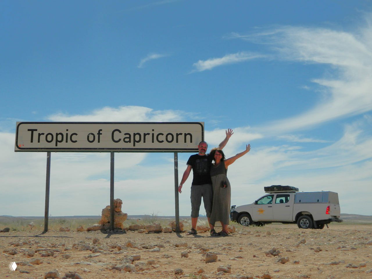 Пара туристов рядом со знаком 'Tropic of Capricorn' на фоне пустынного горизонта и их белый внедорожник, запечатлённый как память о точке маршрута.