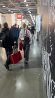Коридор выставочного павильона с посетителями, идущими между стендами; видно людей с сумками и общую активность мероприятия.