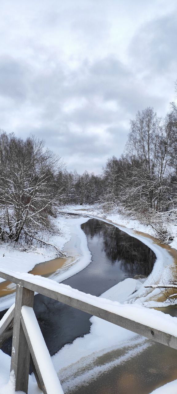Река с частично замёрзшими берегами и вид с перил моста; вокруг снег, деревья и серое зимнее небо, создаётся ощущение тишины и перезагрузки.
