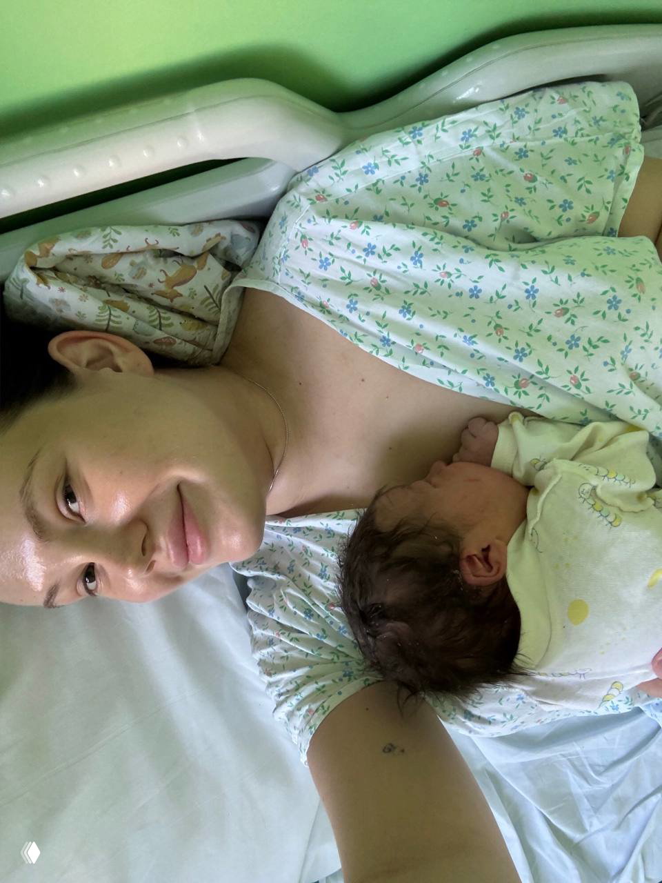 Мама в родильной палате прижимает новорождённого к груди, улыбается в больничной рубашке — спокойная тёплая сцена после родов.