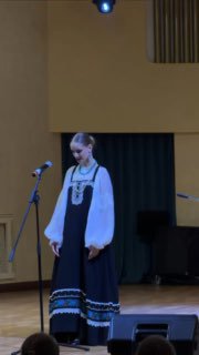 На сцене молодая певица в русском народном костюме исполняет сольное народное пение «Шум берёз», видны микрофон, рояль и затемнённая кулиса.