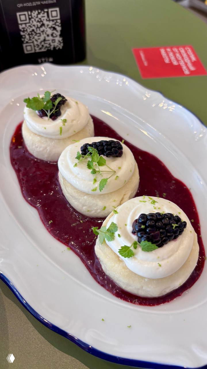 Три нежных сырника на подушке из ежевичного соуса, украшенные ягодой и кремом — подача одного из 15 видов сырников в кофейне Stim, крупный план.