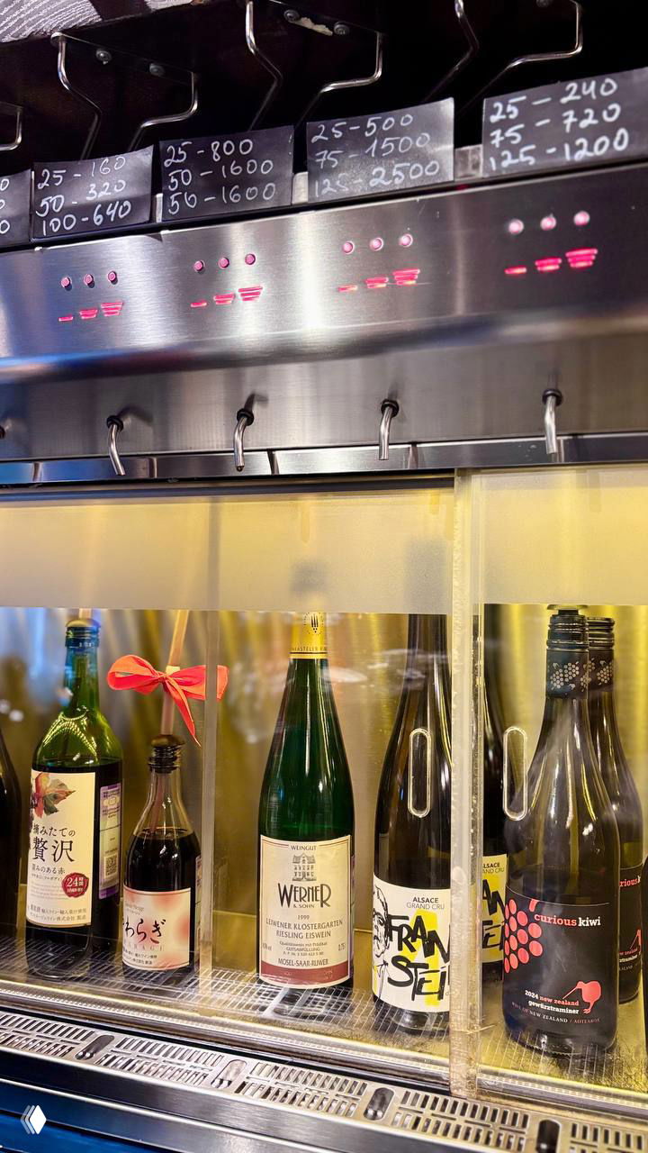 Крупный план форсунок разлива, светодиодных индикаторов и мелованных ценников с указанными объёмами порций и ценами за 25–125 мл в баре.