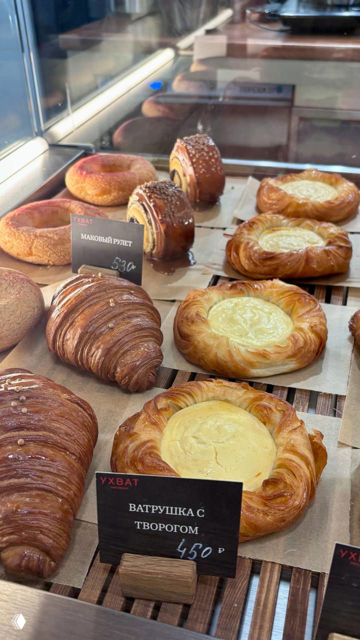 Разнообразная выпечка на подносе: ватрушки с творогом, маковые рулеты и круассаны, аккуратно расставленные в витрине с ценниками.