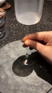 Рука держит бокал на барной стойке во время кофейного омакасе; видны отражения, тень бокала и минималистичная сервировка на столе.