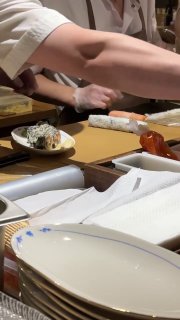 Руки повара на открытой кухне нарезают роллы на деревянной стойке; видно рабочую зону, тарелки и процесс приготовления перед гостями.