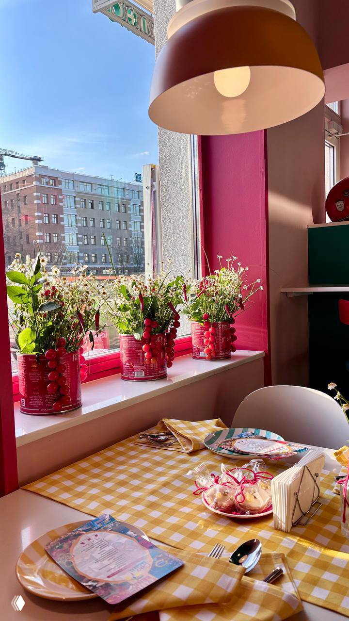 Светлое окно и стол в пиццерии: желтая клетчатая скатерть, банки с растениями на подоконнике и городской вид на улицу Москвы за стеклом.