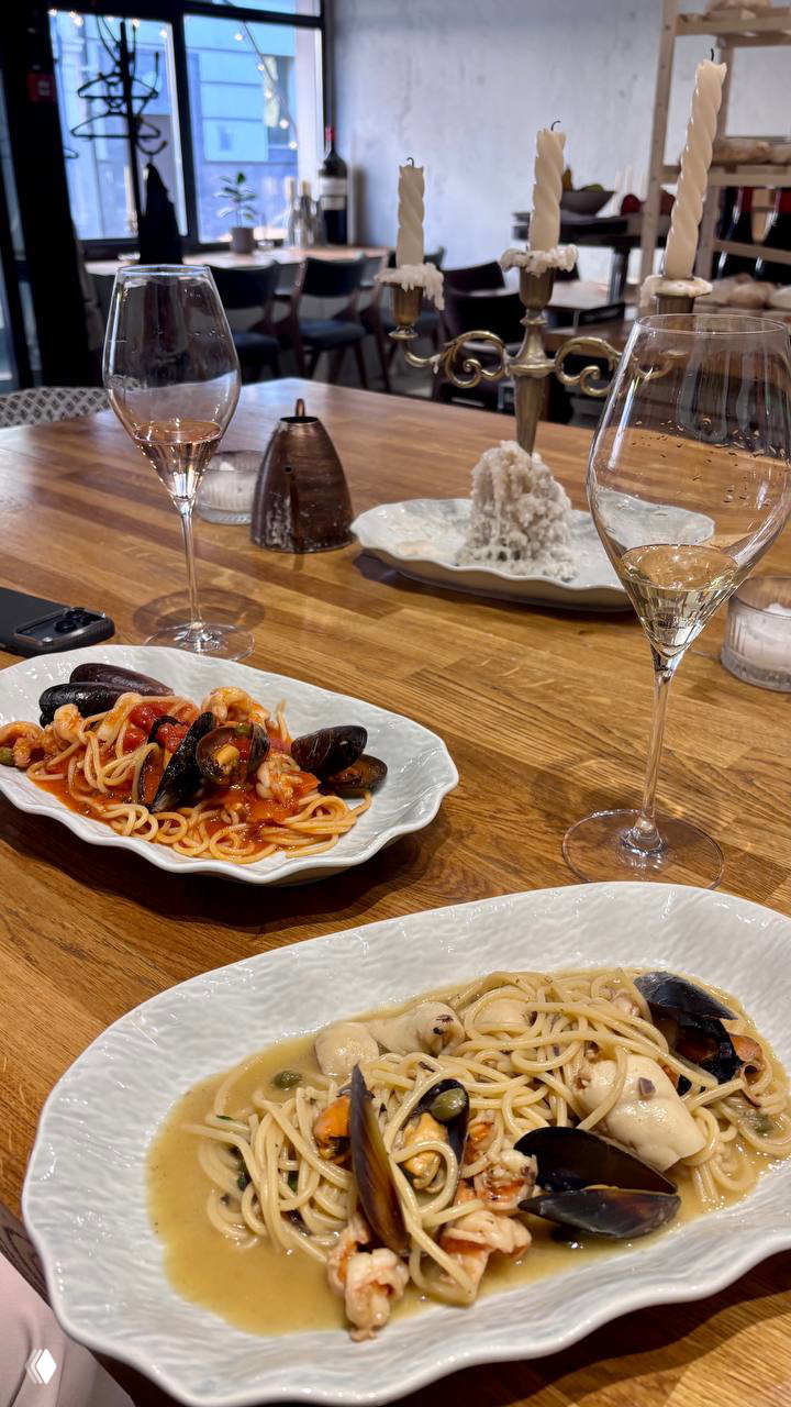 Вид на стол в ресторане: две тарелки с разными пастами, бокалы для вина и свечи на длинной деревянной столешнице в домашнем интерьере