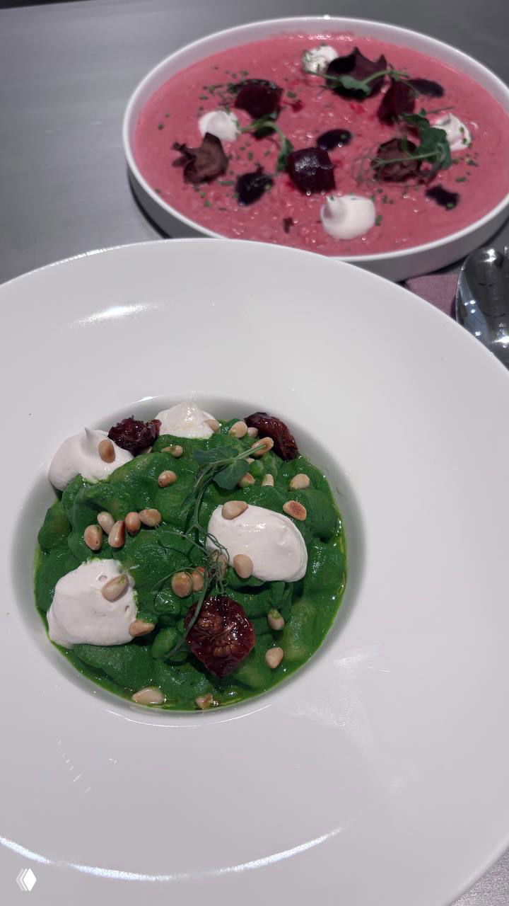 Два блюда на столе в паста-баре Means: зелёная паста и свекольное ризотто рядом, общий вид сервировки, текстуры соусов, порции и цветовые контрасты на белой посуде.