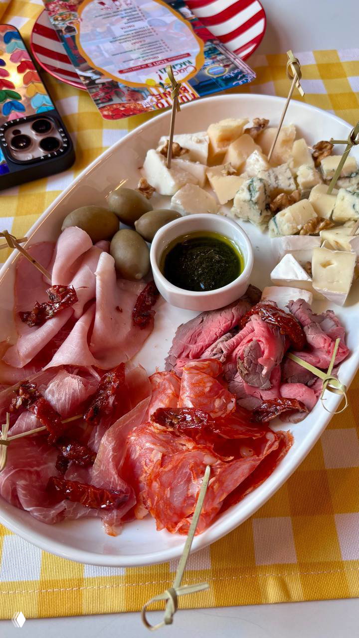 Тарелка антипасто: вяленые томаты, прошутто, ростбиф, оливки и разные сыры на белой овальной подаче на столе.