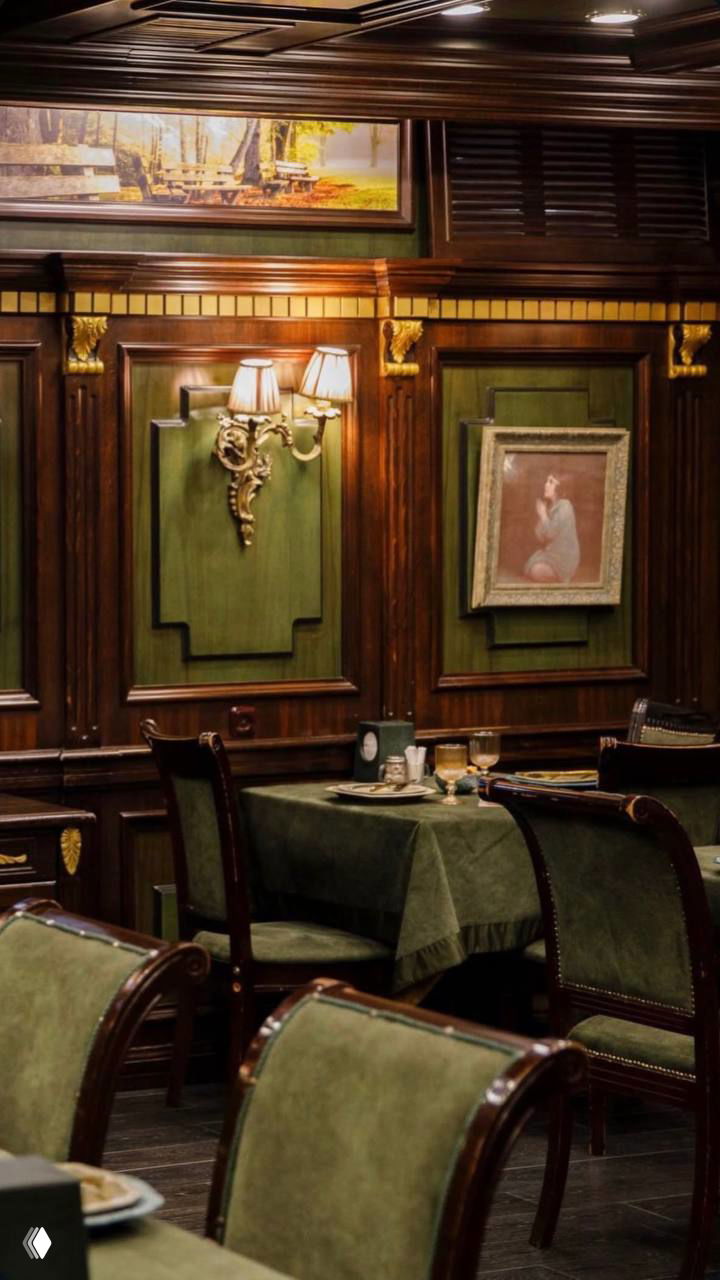 Интерьер ресторана с зелёными обитыми стульями и декоративными деревянными панелями на стенах, мягкое тёплое освещение и картины.