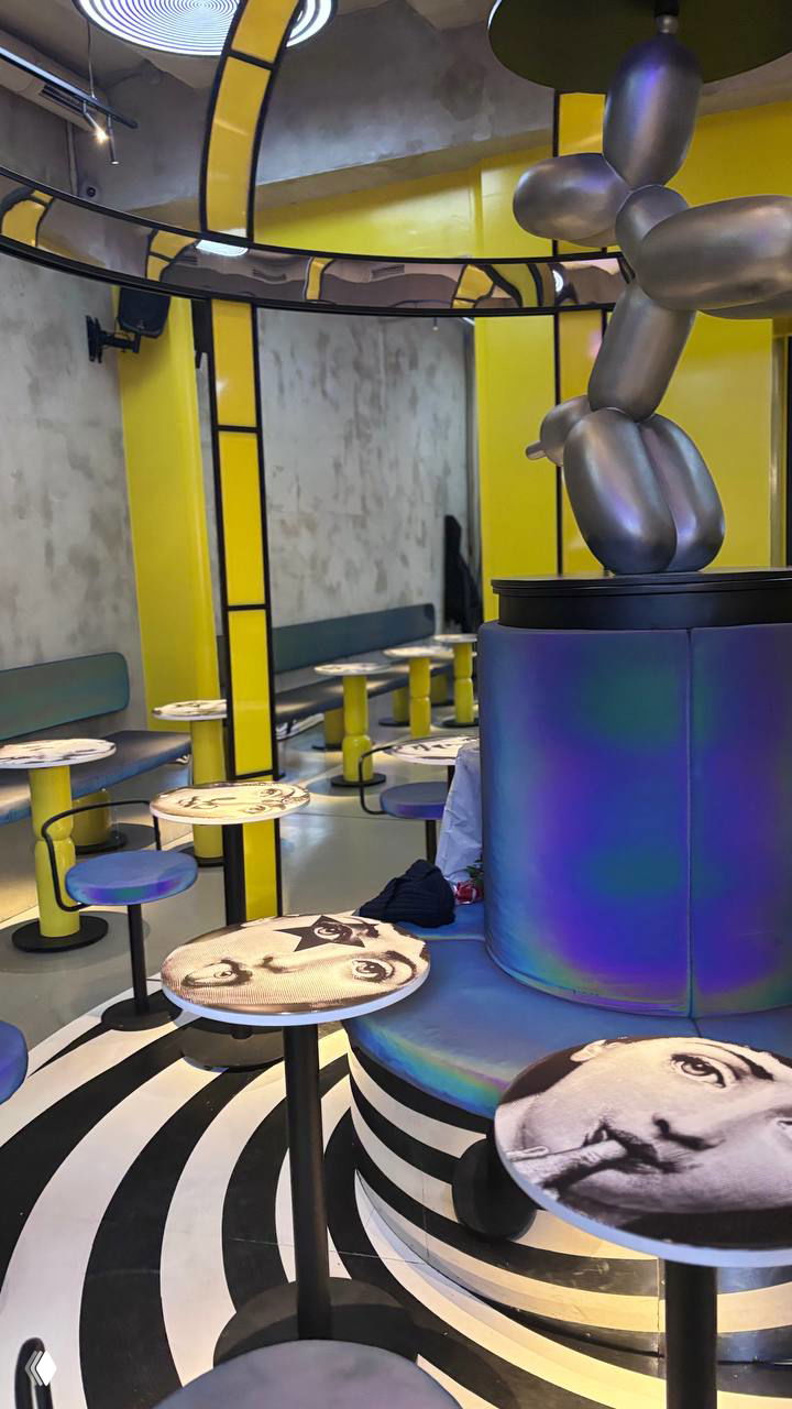 Широкий кадр зала с подчёркнутой чёрно-белой полосатой половой росписью, столиками с лицами и центральным подиумом карусели в жёлто-фиолетовой гамме.