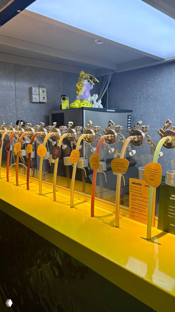 Ряд ярких кранов для напитков на жёлтой стойке, каждый с подписью — часть бара с большим выбором сиропов или лимонадов.