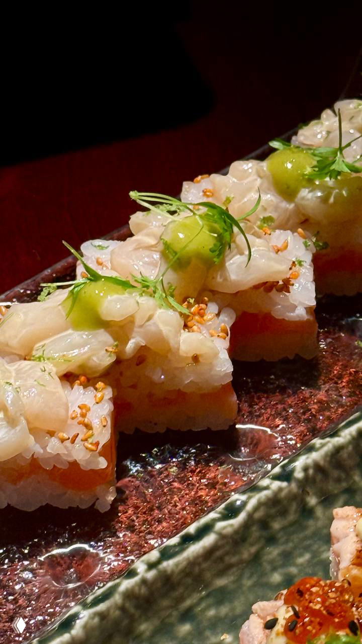 Крупный план квадратных осидзуси с тонкими слайсами рыбы и зелёным соусом: аккуратная текстура риса и декоративные элементы сверху.