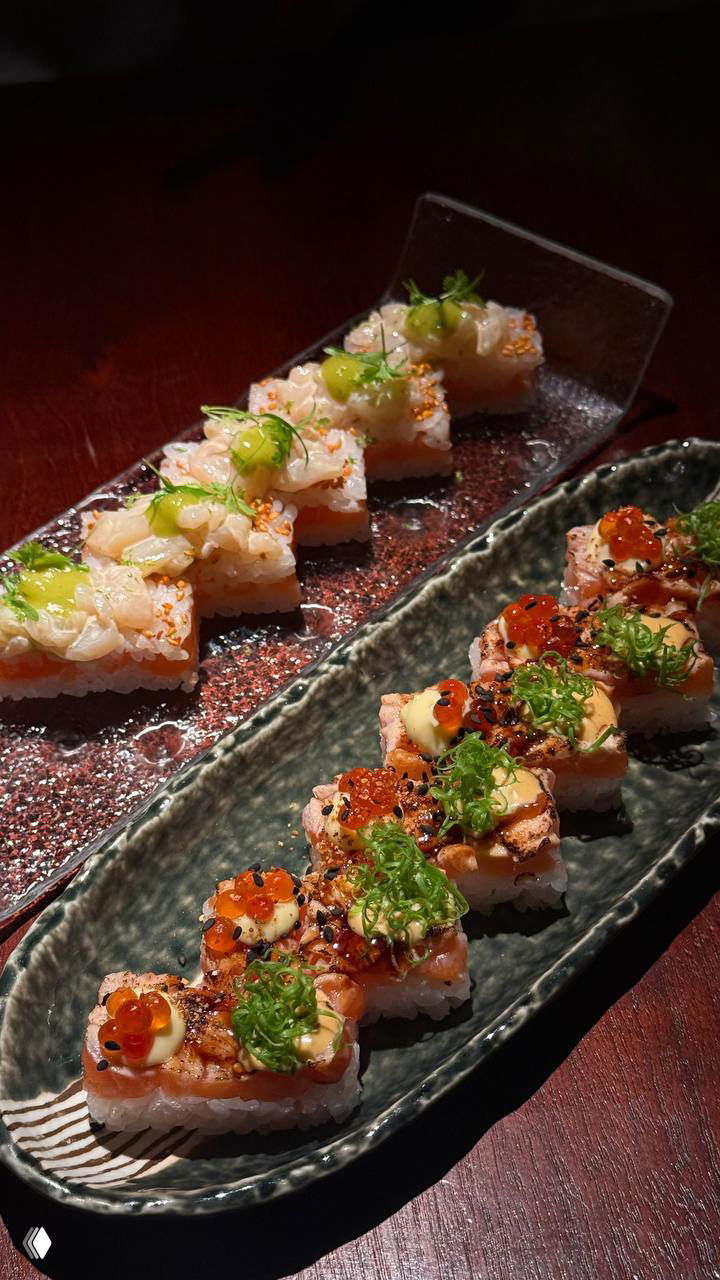 Несколько видов суши и роллов на длинном блюде: яркие соусы, икринки и зелёный лук в декоративной сервировке на тёмной поверхности столика.