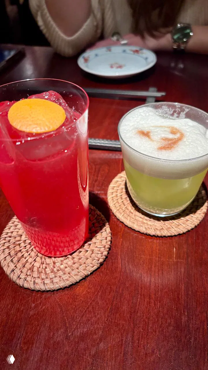 Два коктейля на деревянном столе в Self Edge: ярко-красный и светло-зелёный напитки в стекле, рядом подставки и палочки для суши.