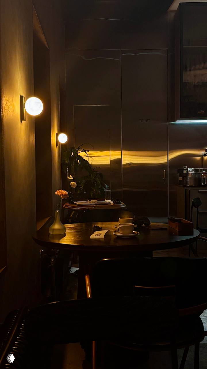 Интерьер Gills при тёплом приглушённом освещении: деревянный стол, чашки, ваза с цветком и отражающая металлическая стена на заднем плане