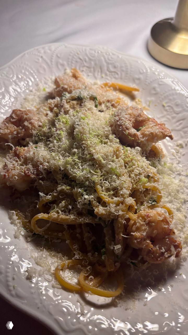 Тарелка с пастой и жареными креветками, щедро посыпанная тёртым сыром, снята под настольной лампой на белой скатерти в интерьере бара Louis.