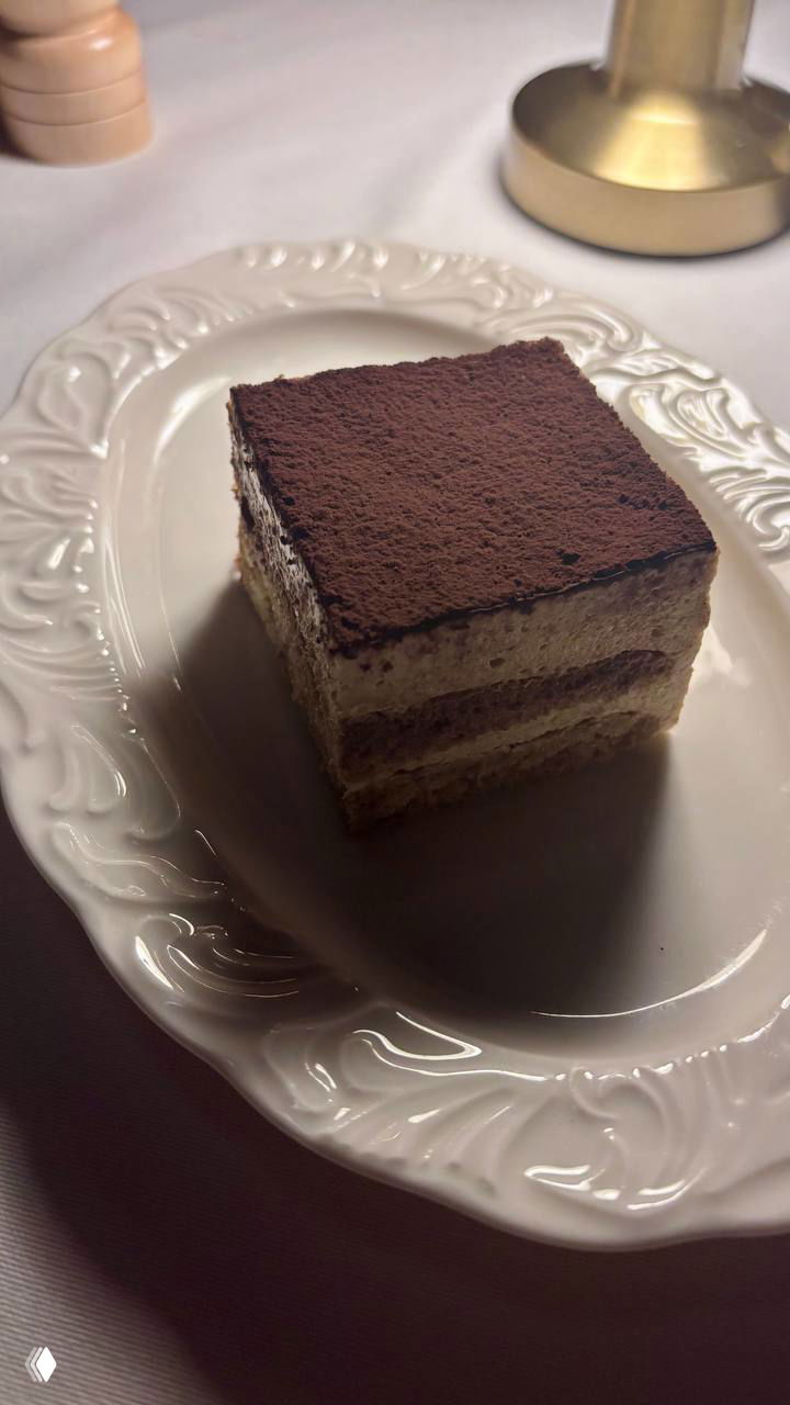 Кусок тирамису на узорной белой тарелке, сверху какао-порошок; десерт снят при боковом свете в ресторане Louis, подчёркивающем текстуру и форму.