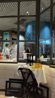 Интерьер La Bottega Siciliana: открытая кухня и печь, повара готовят за стеклом, стол с бутылками масла и сервировкой, вид из зала на рабочую зону.