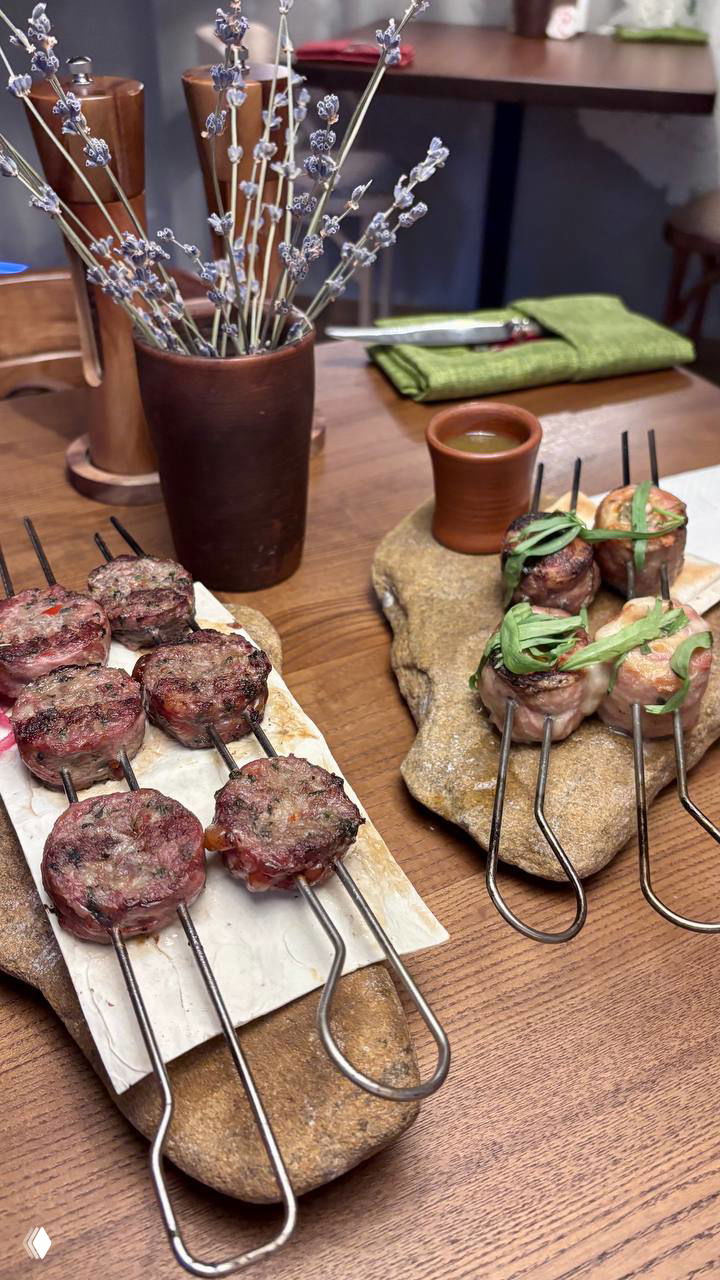Две деревянные подставки с металлическими шампурами и мини‑шашлыками, украшенные зеленью и поданные на каменных подложках в интерьере кафе.