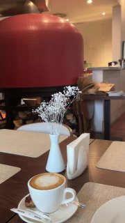 Чашка капучино на столе в интерьере ресторана: маленькая ваза с сухоцветами, вид на большой красный купол печи и обслуживающую зону.