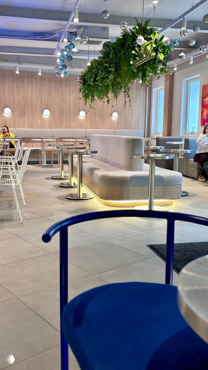 Светлое и просторное пространство зала с мягкими диванами и зеленой растительной декорацией — интерьер, похожий на кафе, где подают бургеры.