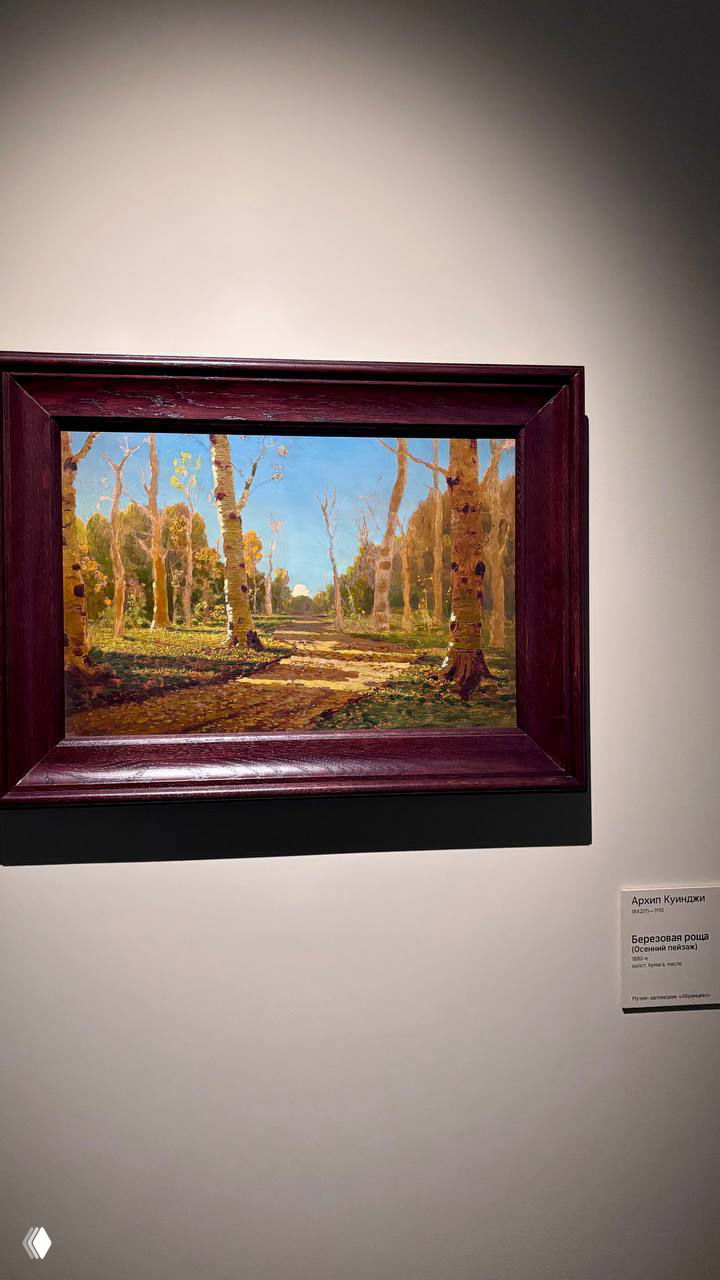 Небольшая живописная работа в тёмной рамке на белой стене; изображён солнечный лесной просёлок с берёзами и светом, подпись автора на табличке.