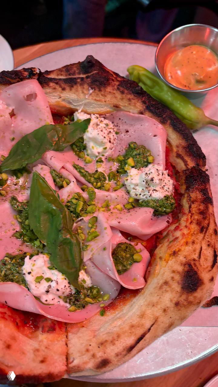 Крупный план пиццы с ломтиками мяса (мортаделла/хамон), рикоттой, песто и дроблёным фисташком на пышном обугленном бортике.