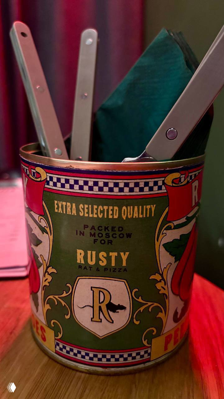 Декоративная банка с логотипом Rusty Rat & Pizza, в ней стоящие салфетки и столовые приборы — деталь интерьера ресторана.